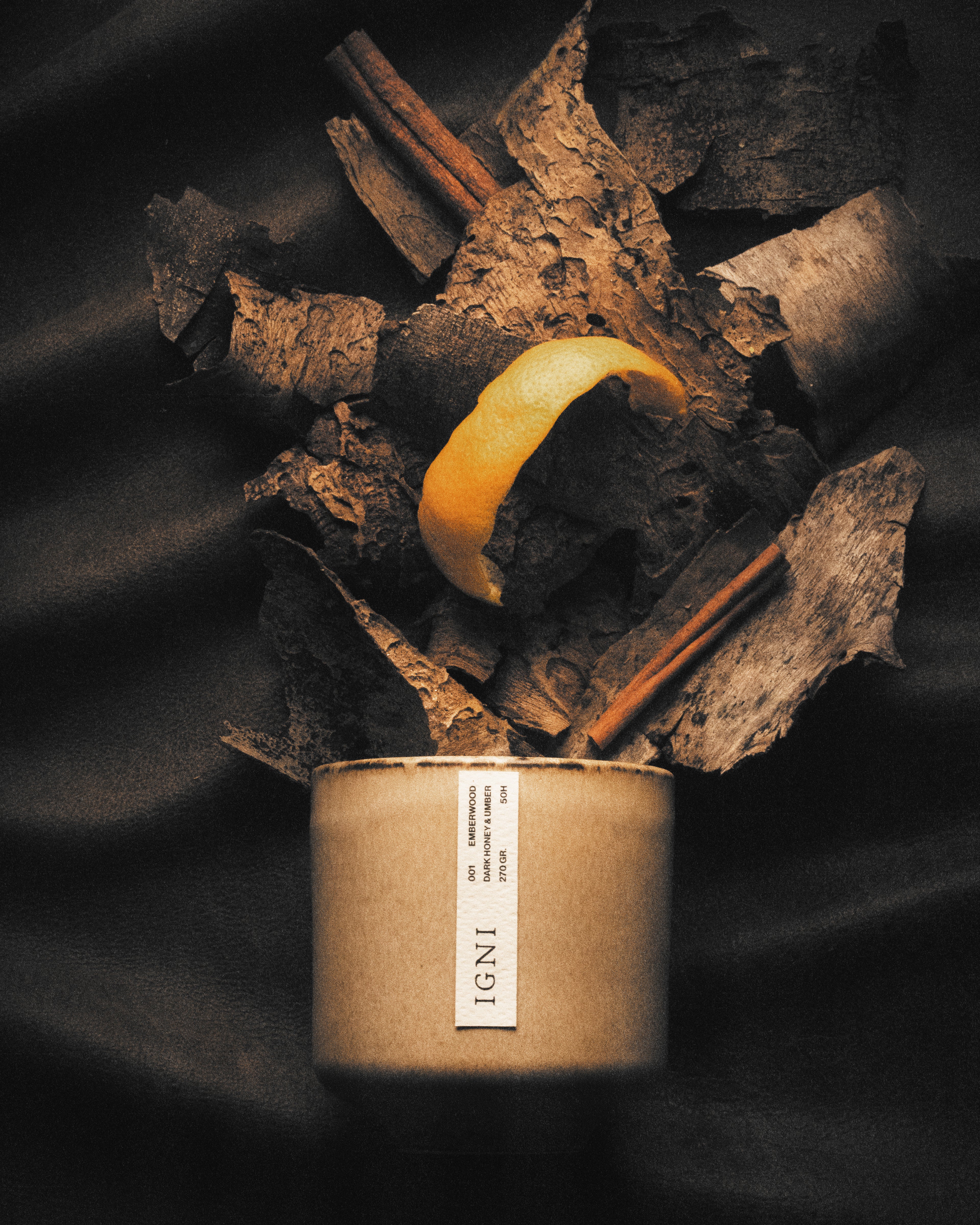 IGNI Emberwood Dark Honey & Umber luxury scented candle in a beige ceramic vessel, styled with layered wood bark, cinnamon sticks, citrus peel, and dark moody textures.