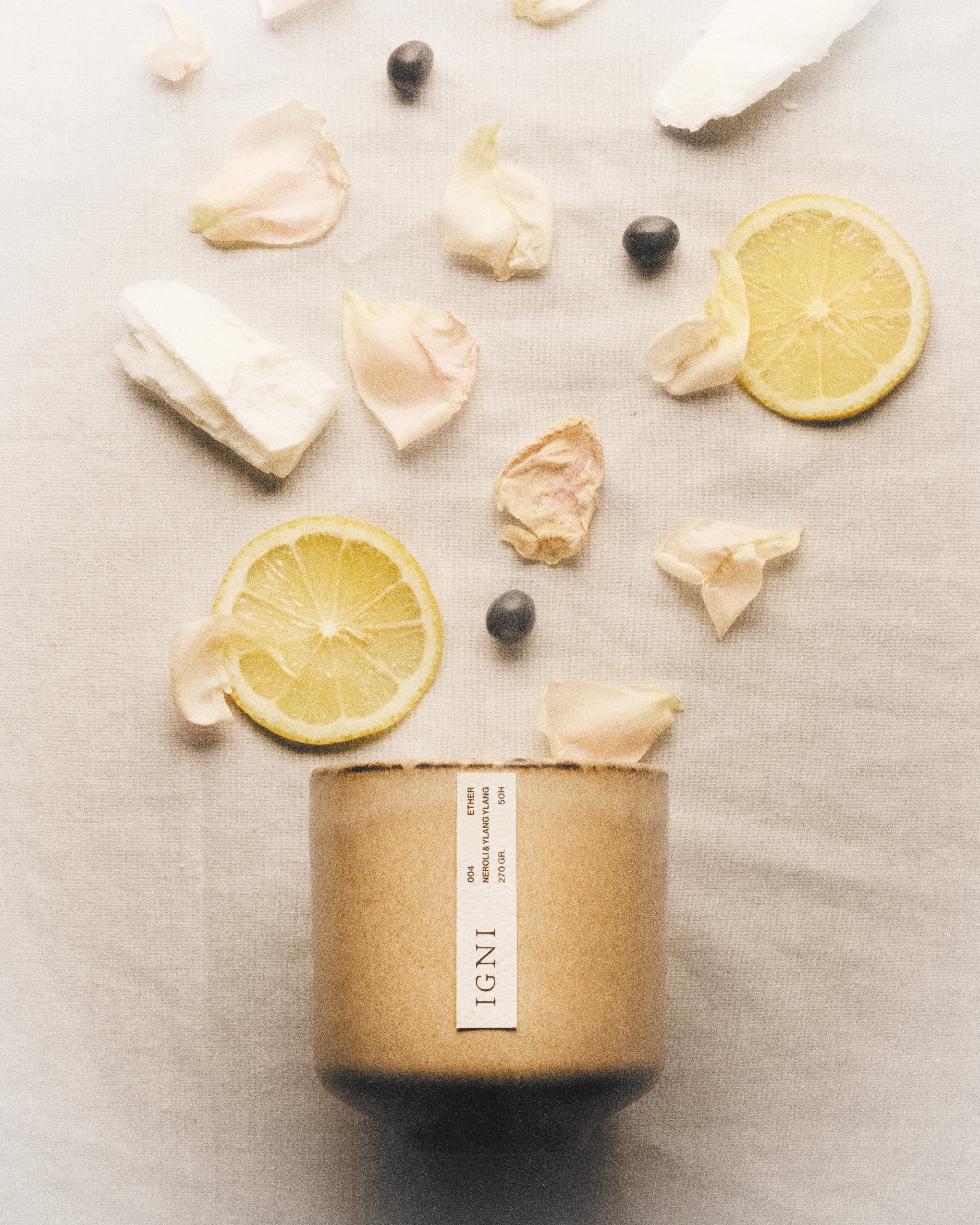 IGNI Ether Neroli & Ylang Ylang luxury scented candle in a beige ceramic vessel, styled with lemon slices, pale flower petals, dark seeds, and soft linen textures.