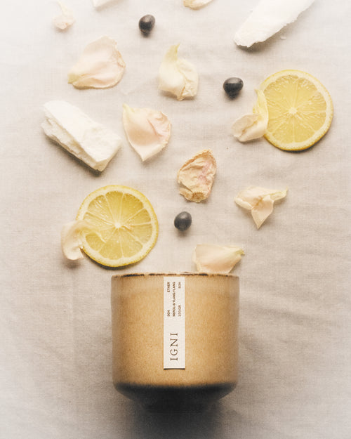 IGNI Ether Neroli & Ylang Ylang luxury scented candle in a beige ceramic vessel, styled with lemon slices, pale flower petals, dark seeds, and soft linen textures.
