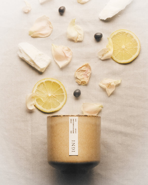 IGNI Ether Neroli & Ylang Ylang luxury scented candle in a beige ceramic vessel, styled with lemon slices, pale flower petals, dark seeds, and soft linen textures.