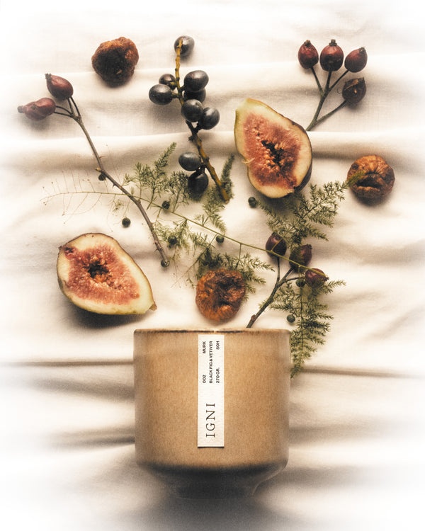 IGNI Murk Black Fig & Vetiver luxury scented candle in a beige ceramic vessel, styled with fresh figs, dried botanicals, dark berries, and soft fabric textures.
