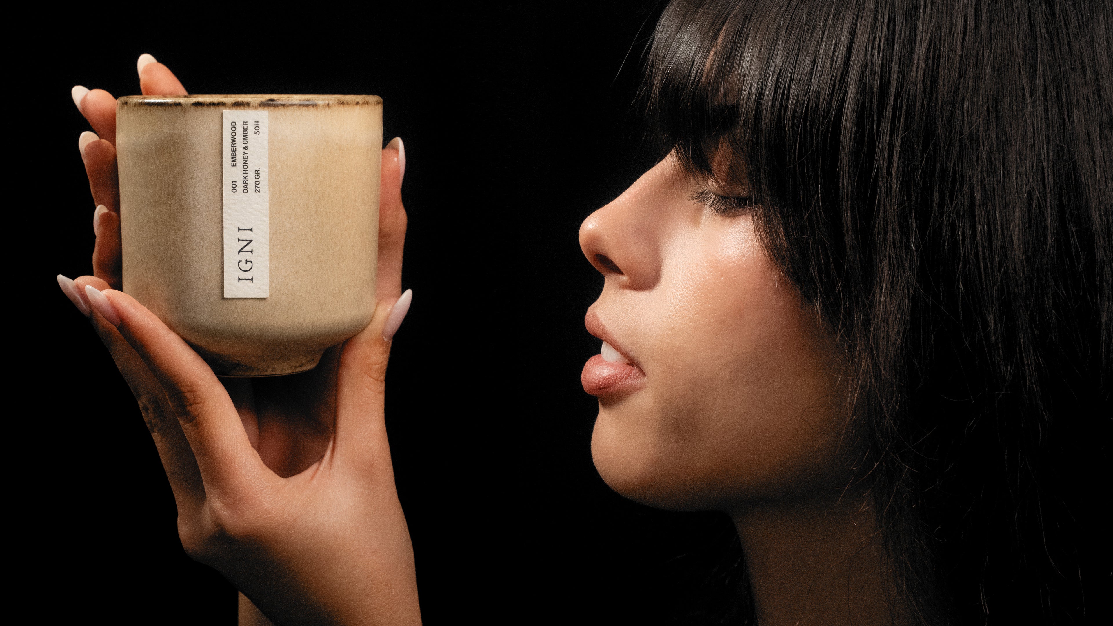 Close-up side portrait of a model holding a luxury scented IGNI candle in a beige ceramic vessel against a black background with warm studio lighting.