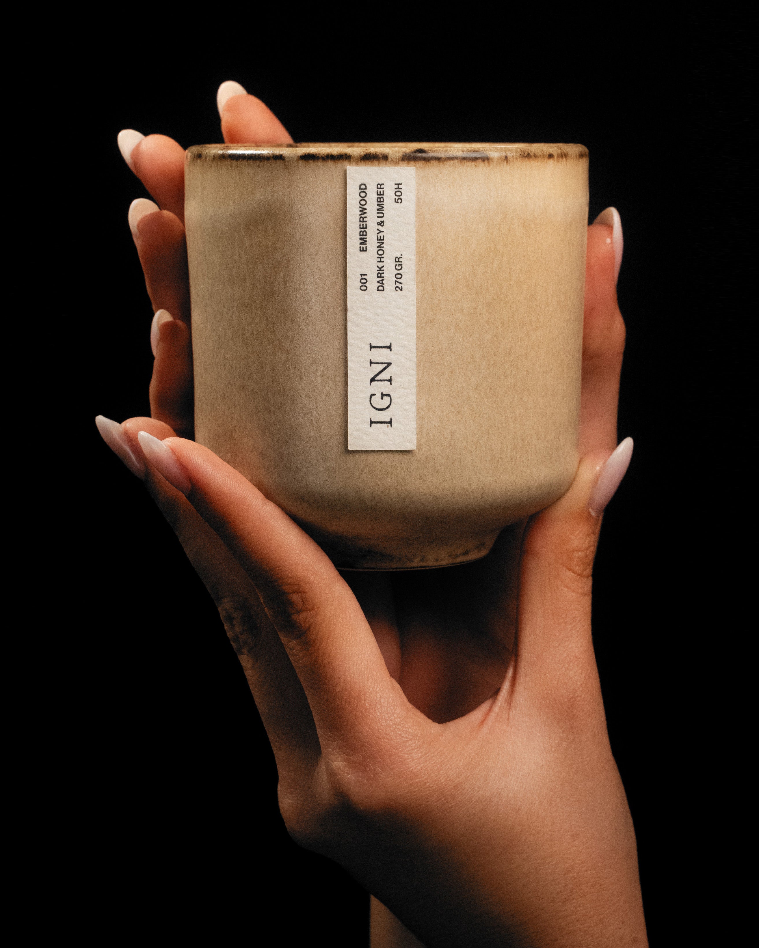 Hands holding a luxury IGNI Emberwood scented candle in a beige ceramic vessel, featuring a Dark Honey & Umber fragrance label against a black background.