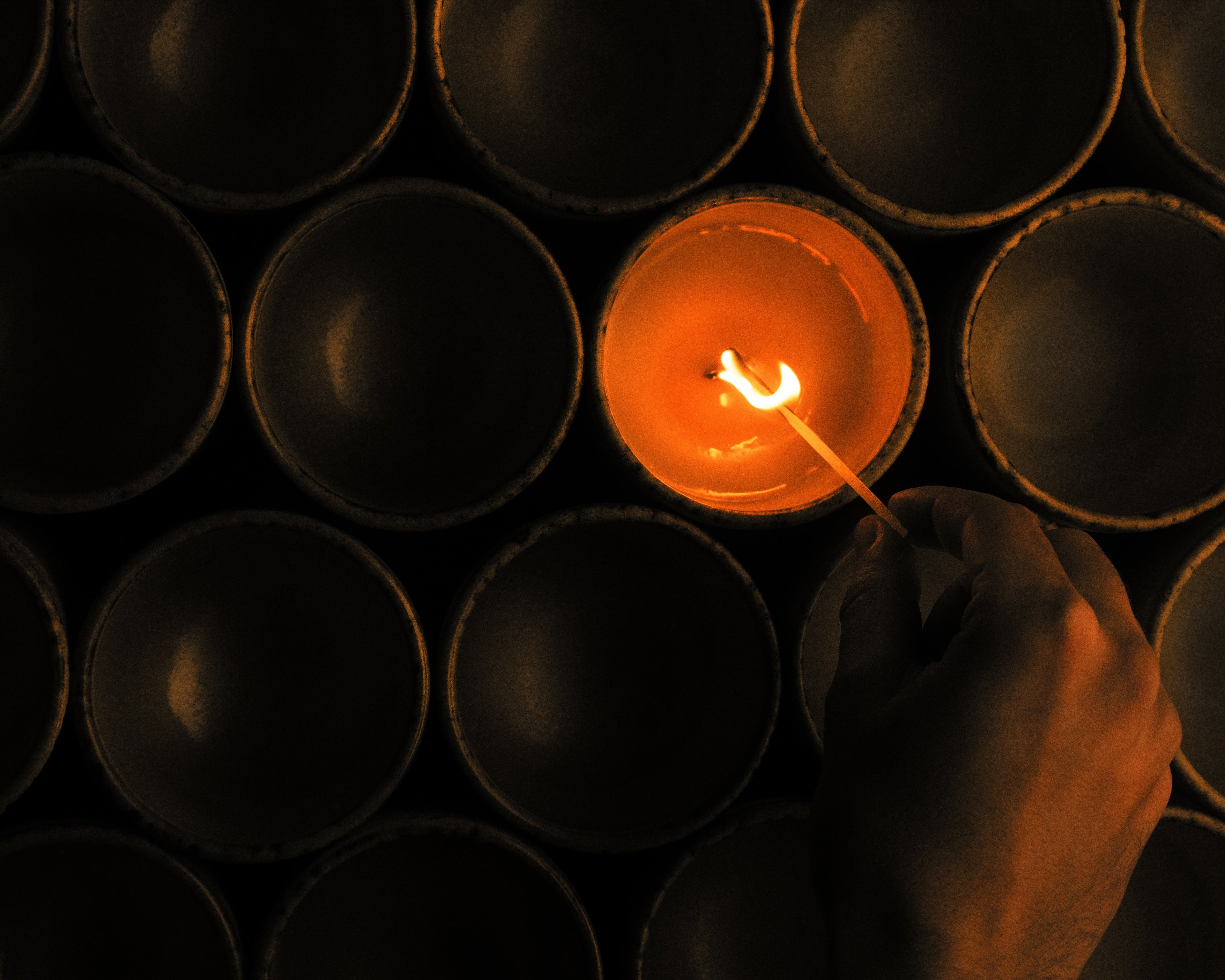 Close-up of a luxury scented candle being lit with a match, glowing warm amber among dark ceramic vessels for an atmospheric artisanal candle-making scene.