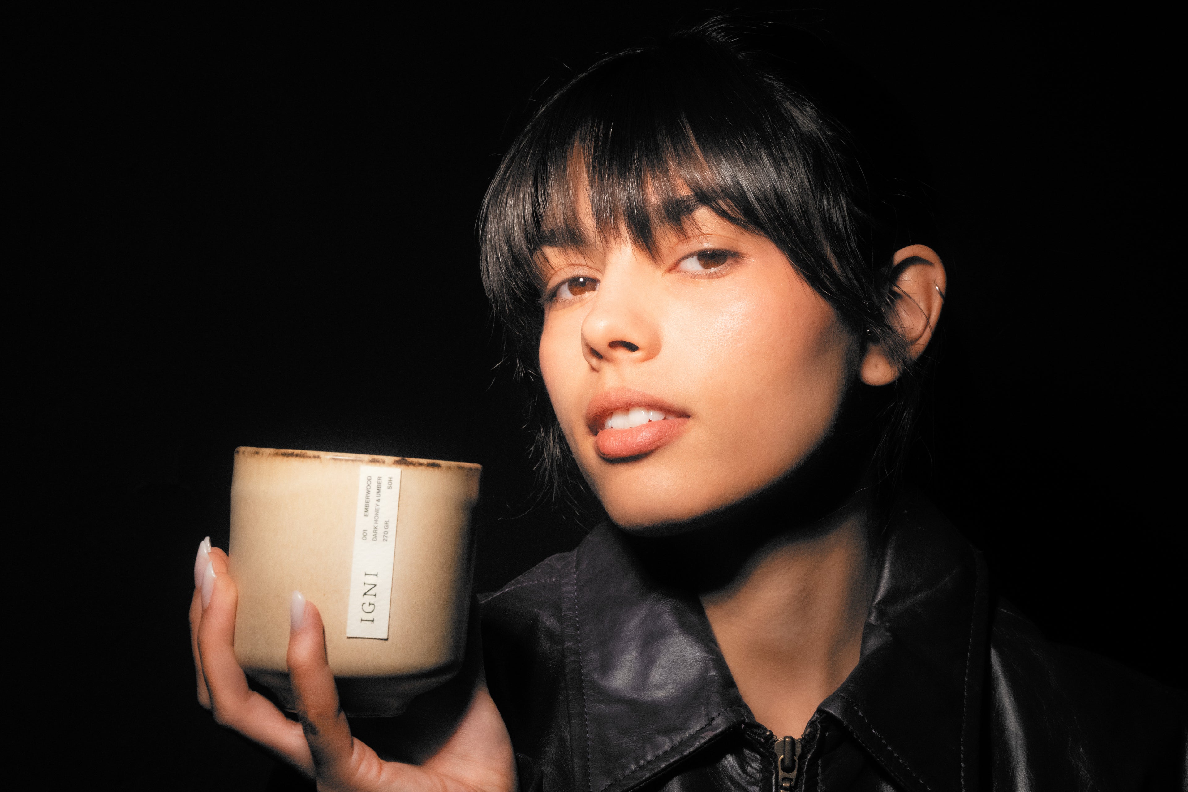 Portrait of a model holding an IGNI Emberwood luxury scented candle in a beige ceramic vessel, set against a black background with warm dramatic lighting.