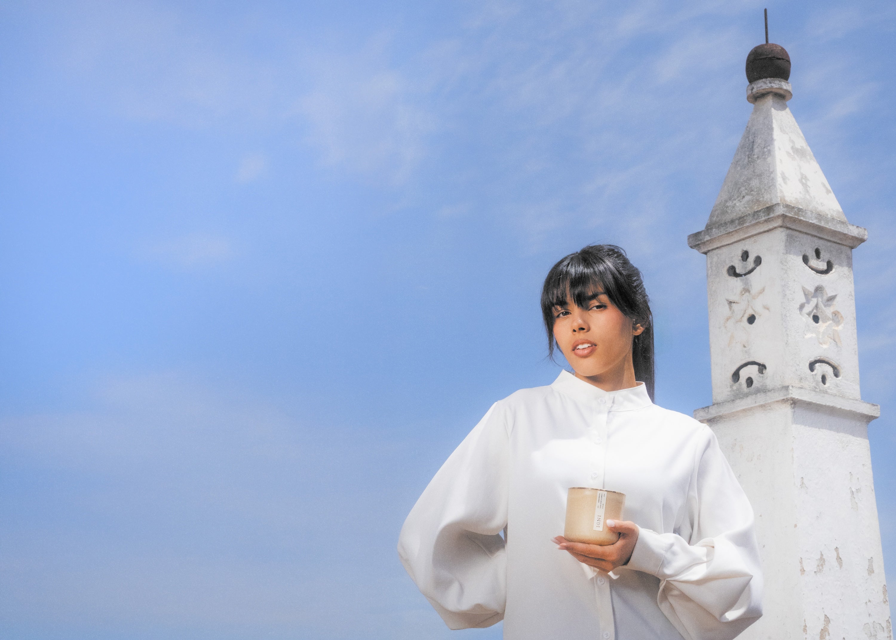 Model in a white blouse holding an IGNI luxury scented candle in a beige ceramic vessel, standing outdoors under a blue sky beside a white Traditional Algarve Chimney.